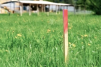 Vermessungsleistung - Grob- und Feinabsteckung Das Bild zeigt einen Vermessungspflock auf einer Wiese vor einer Häuserkulisse