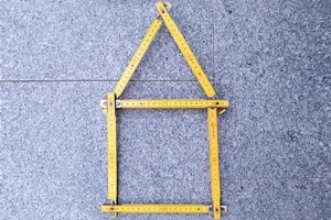 Vermessungsleistung - Gebäudeeinmessung Das Bild zeigt ein Haus gebaut aus einem gelben Zollstock vor einem Steinhintergrund